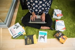 A woman sitting on the floor using a laptop, with tech books around her, symbolizing women in tech