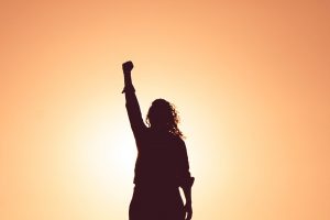 Silhouette of a women raising her fist in the air in front of an orange sky, representing women's empowerment