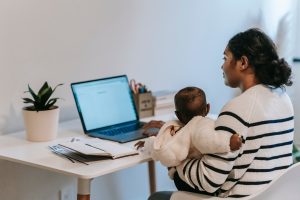 A woman holds a baby while working on a laptop, representing The Mom Project