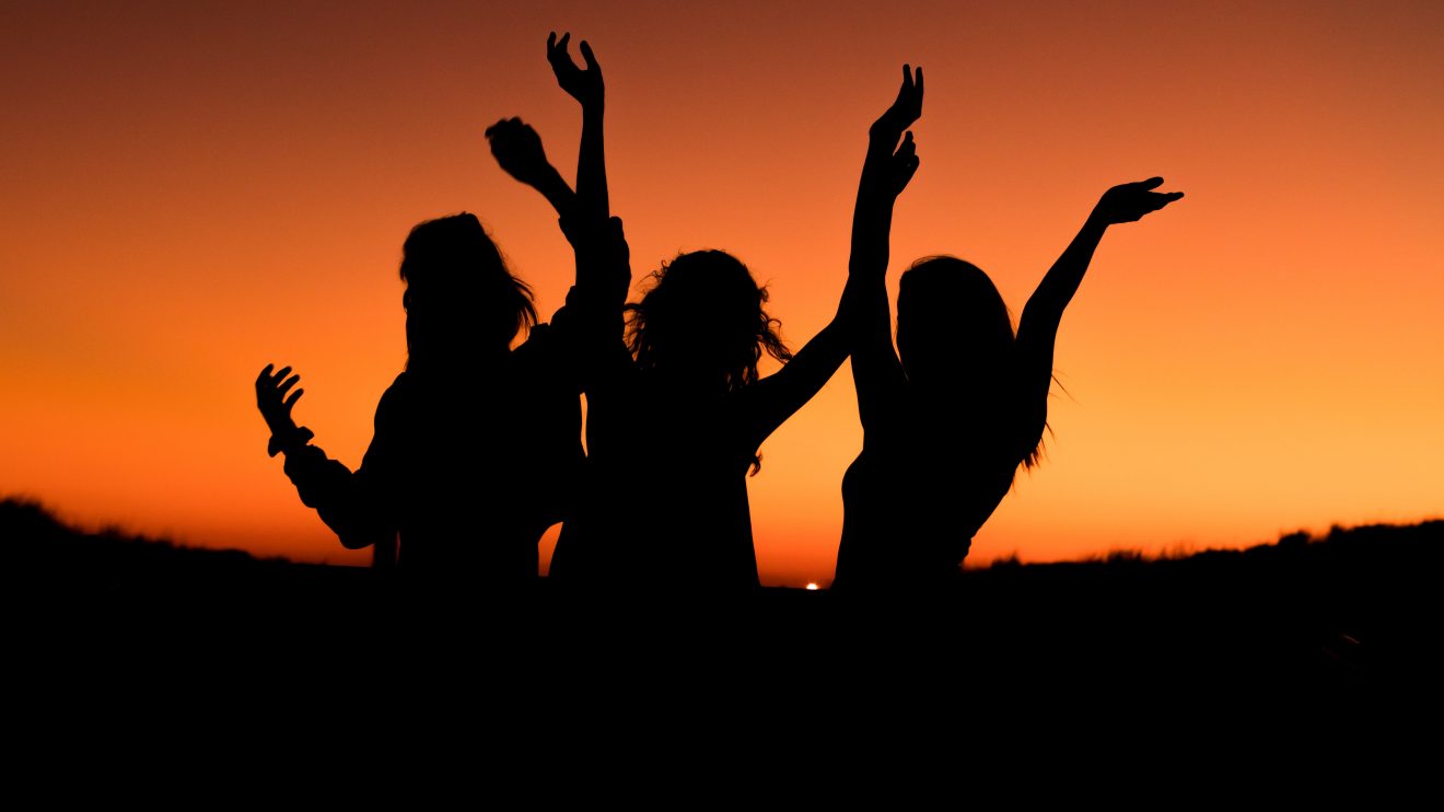 Three silhouettes of women with their arms up in front of an orange sunset