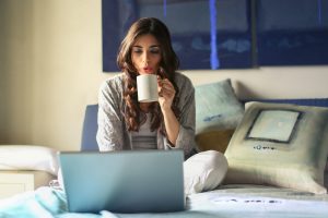 Woman in bed on laptop, sipping coffee from a mug, representing post-pandemic hybrid work