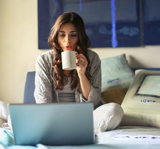 Woman in bed on laptop, sipping coffee from a mug, representing post-pandemic hybrid work