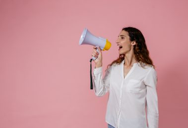 Woman shouting into a speakerphone against a pink background, representing "Women at the Helm" speaker series