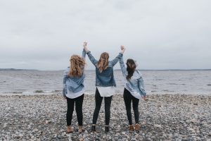 Three women at the ocean holding their hands up together, representing OUTFRONT Media "Lifted Up" campaign