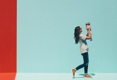 A mother in a striped shirt holds her baby up playfully against a colorful background split into red and blue sections. This image illustrates the impact of life and family events on entrepreneurial success.
